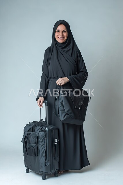 الانطلاق لقضاء رحلة صيفية ممتعة, الشعور بالسعادة والفرح، السياحة في إجازة الصيف, بورتريه لشابة عربية خليجية سعودية محجبة ترتدي العباءة السوداء تمسك حقيبة السفر تنظر إلى الكاميرا بإيماءات الحماس, صورة الجسم بالطول الكامل, خلفية رماديه