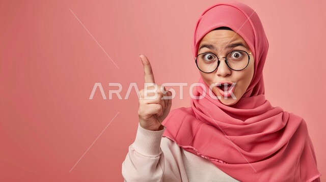 ايماءات تدل على الذهول لسماع خبر مفاجئ، الصدمة والتعجب من حصول أمر ما، بورتريه مقرب لشابة عربية خليجية سعودية محجبة تنظر إلى الكاميرا بتعابير الدهشة والخوف تشير بأصبع السبابة الى مكان ما، خلفية وردية
