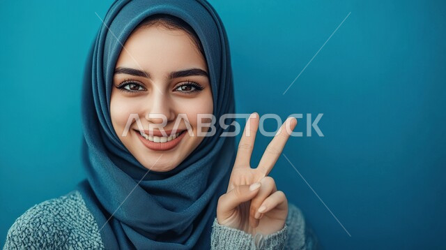 مفهوم الفرح بالفوز، النجاح وتحقيق الأهداف، تعابير السرور والسعادة، بورتريه مقرب لامرأة عربية خليجية سعودية محجبة مبتسمة محجبة ترفع يديها بعلامة النصر تنظر الى الكاميرا بايماءات الحماس، خلفية ملونة