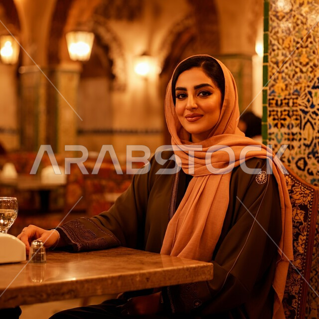 مطاعم و كافيهات المملكة العربية السعودية, قضاء أوقات ممتعة في المطعم, امرأة عربية خليجية سعودية مبتسمة محجبة ترتدي العباءة السوداء تجلس على المائدة و تنظر إلى الكاميرا بإيماءات السعادة و السرور