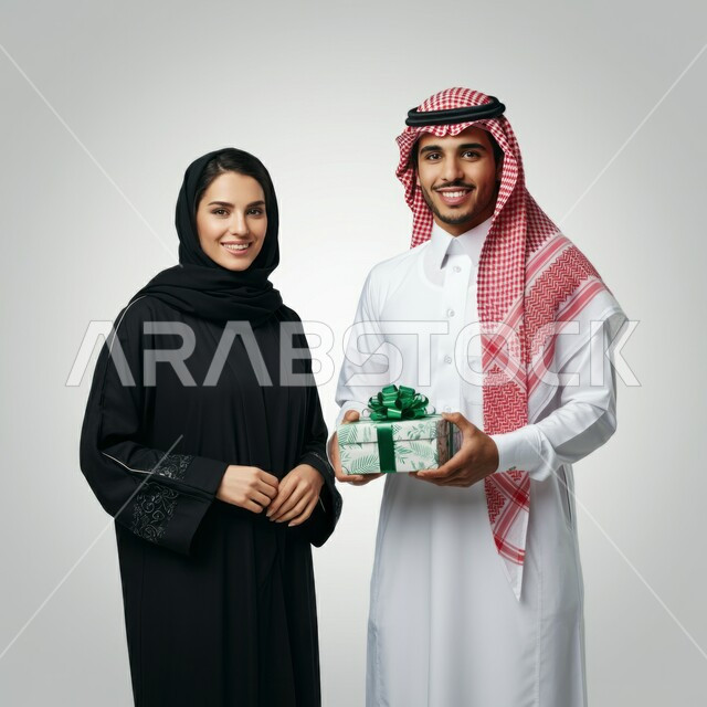 الألفة والمحبة بين الزوجين, مشاركة وتقديم الهدايا في الأعياد والمناسبات العائلية, رجل عربي خليجي سعودي يرتدي الثوب التقليدي والشماغ يقدم لزوجته هدية بمناسبة ذكرى يوم الزواج,    مفهوم الترابط الأسري، النظر إلى الكاميرا بإيماءات السعادة والسرور، خلفية بيضاء