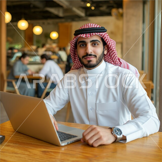 استخدام الأجهزة التقنية الحديثة في الحياة اليومية, قضاء الأوقات الممتعة في المطعم, شاب عربي خليجي سعودي مبتسم يرتدي الثوب التقليدي و الشماغ يجلس على الطاولة و يستعمل جهاز الحاسوب المحمول, النظر إلى الكاميرا بإيماءات السعادة و السرور