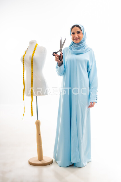 ورشة عمل تصميم ازياء، اختراع وابتكار موديلات جديدة وعصرية، أهمية المرأة في مجال الخياطة، مهن وأعمال فنية نسائية، بورتريه لمصممة عربية خليجية عمانية مبتسمة ترتدي عباءة زرقاء تحمل بيدها مقص، فنون ومهارات تفصيل الملابس النسائية، مفهوم عالم الأزياء والموضة