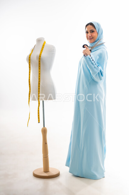 ورشة عمل تصميم ازياء، اختراع وابتكار موديلات جديدة وعصرية، أهمية المرأة في مجال الخياطة، مهن وأعمال فنية نسائية، بورتريه لمصممة عربية خليجية عمانية مبتسمة ترتدي عباءة زرقاء تحمل بيدها مقص، فنون ومهارات تفصيل الملابس النسائية، مفهوم عالم الأزياء والموضة