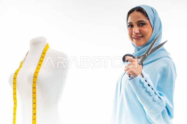 Fashion design workshop, invention and innovation of new and modern models, the importance of women in the field of sewing, women's professions and artistic works, portrait of a smiling Gulf Arab Omani designer wearing a blue abaya and holding scissors in her hand, arts and skills of tailoring women's clothing, the concept of the world of fashion and style