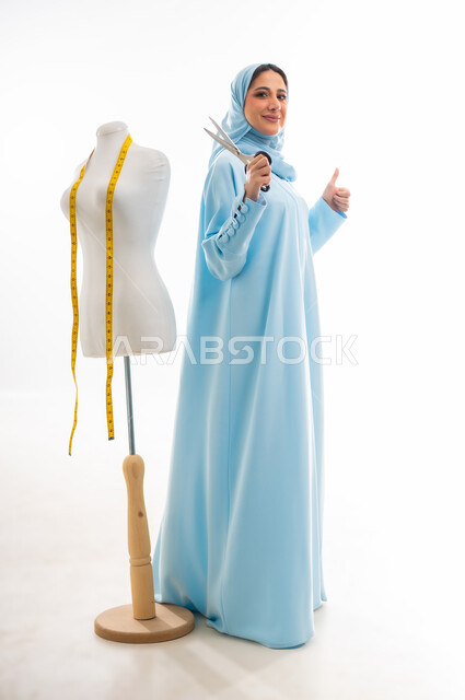 Fashion design workshop, invention and innovation of new and modern models, the importance of women in the field of sewing, women's professions and artistic works, portrait of a smiling Gulf Arab Omani designer wearing a blue abaya and holding scissors in her hand, arts and skills of tailoring women's clothing, the concept of the world of fashion and style