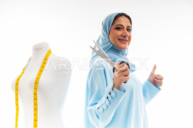 ورشة عمل تصميم ازياء، اختراع وابتكار موديلات جديدة وعصرية، أهمية المرأة في مجال الخياطة، مهن وأعمال فنية نسائية، بورتريه لمصممة عربية خليجية عمانية مبتسمة ترتدي عباءة زرقاء تحمل بيدها مقص، فنون ومهارات تفصيل الملابس النسائية، مفهوم عالم الأزياء والموضة