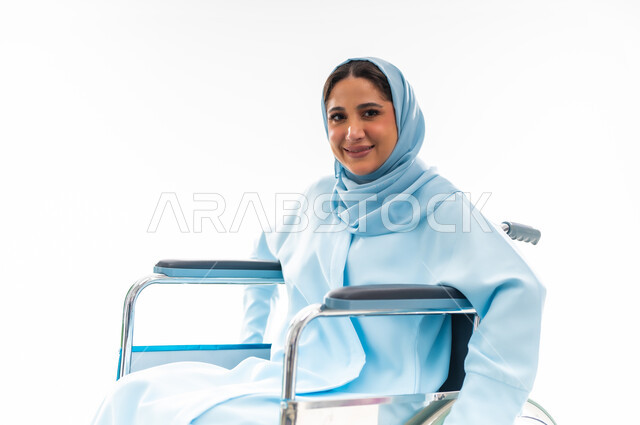 The concept of optimism and love of life, caring for people with disabilities and providing for their needs, a portrait of a smiling Gulf Arab Omani woman wearing a hijab and an abaya with special needs sitting in a wheelchair, providing devices and equipment that help with movement and mobility, white background