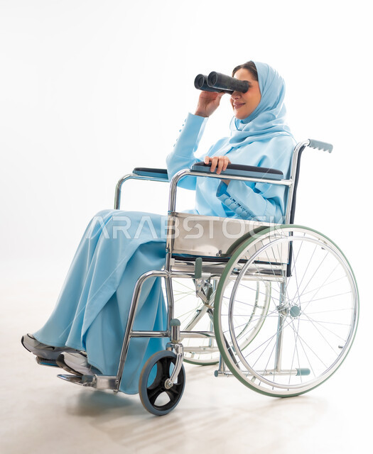 مفهوم التفاؤل وحب الحياة، العنايه بأصحاب الهمم وتأمين احتياجاتهم، بورتريه لإمرأة  عربية خليجية عمانية مبتسمة ترتدي الحجاب والعباءة من ذوي الاحتياجات الخاصه تجلس على كرسي متحرك، توفير اجهزة ومعدات تساعد بالحركة والتنقل، خلفيه بيضاء