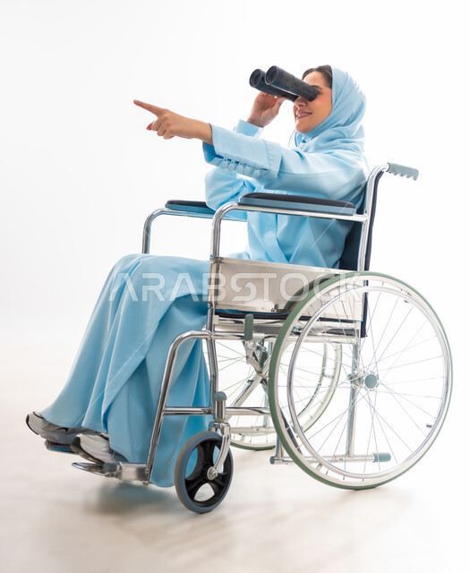 مفهوم التفاؤل وحب الحياة، العنايه بأصحاب الهمم وتأمين احتياجاتهم، بورتريه لإمرأة  عربية خليجية عمانية مبتسمة ترتدي الحجاب والعباءة من ذوي الاحتياجات الخاصه تجلس على كرسي متحرك، توفير اجهزة ومعدات تساعد بالحركة والتنقل، خلفيه بيضاء