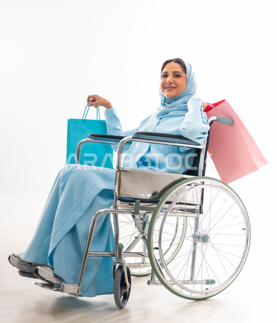 مفهوم التفاؤل وحب الحياة، العنايه بأصحاب الهمم وتأمين احتياجاتهم، بورتريه لإمرأة  عربية خليجية عمانية مبتسمة ترتدي الحجاب والعباءة من ذوي الاحتياجات الخاصه تجلس على كرسي متحرك، توفير اجهزة ومعدات تساعد بالحركة والتنقل، خلفيه بيضاء