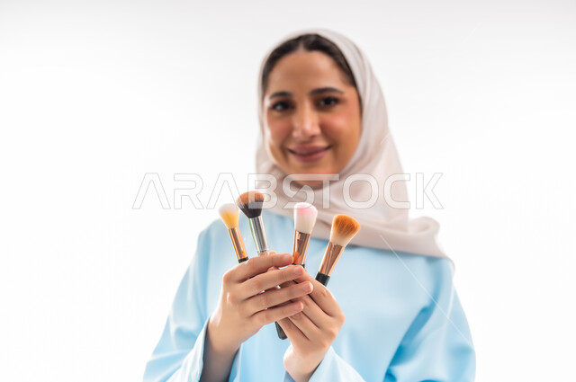 طرق مبتكرة للدعاية والاعلان، الأنوثة والاهتمام بالجمال والبشرة والمظهر الخارجي, خبيرة لمستحضرات التجميل النسائية، بورتريه مقرب لإمرأة عربية خليجية عمانية ترتدي العباءة والحجاب تحمل بيدها أدوات خاصة بالمكياج، التسويق لمنتجات الميك اب، خلفية بيضاء