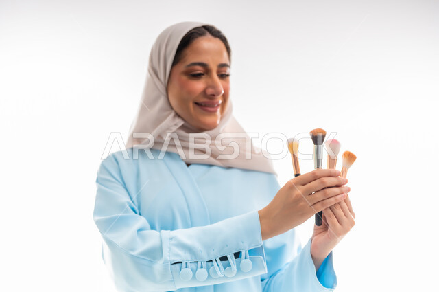 طرق مبتكرة للدعاية والاعلان، الأنوثة والاهتمام بالجمال والبشرة والمظهر الخارجي, خبيرة لمستحضرات التجميل النسائية، بورتريه مقرب لإمرأة عربية خليجية عمانية ترتدي العباءة والحجاب تحمل بيدها أدوات خاصة بالمكياج، التسويق لمنتجات الميك اب، خلفية بيضاء