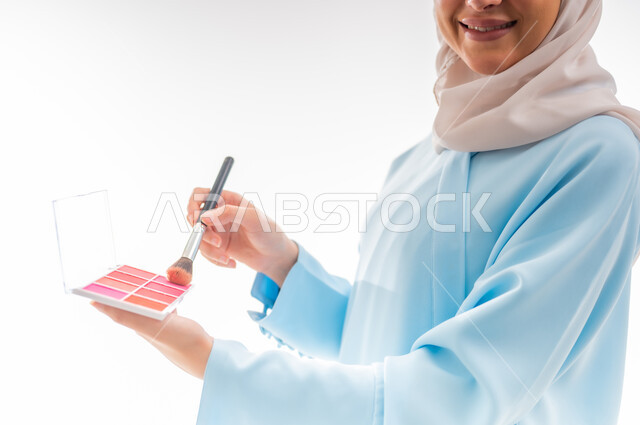 الأنوثة والاهتمام بالجمال والبشرة والمظهر الخارجي, مفهوم الدعاية والإعلان، خبيرة لمستحضرات التجميل النسائية، بورتريه مقرب لإمرأة عربية خليجية عمانية ترتدي العباءة والحجاب تقوم بوضع أحمر الخدود، التسويق لمنتجات الميك اب، خلفية بيضاء