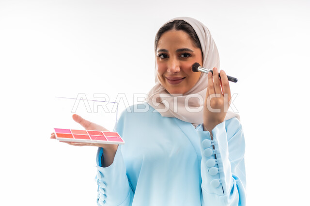 الأنوثة والاهتمام بالجمال والبشرة والمظهر الخارجي, مفهوم الدعاية والإعلان، خبيرة لمستحضرات التجميل النسائية، بورتريه مقرب لإمرأة عربية خليجية عمانية ترتدي العباءة والحجاب تقوم بوضع أحمر الخدود، التسويق لمنتجات الميك اب، خلفية بيضاء