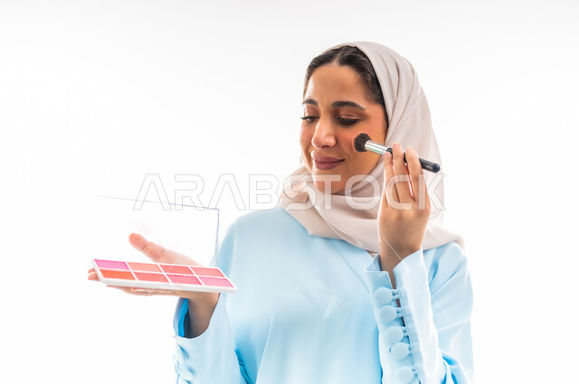 الأنوثة والاهتمام بالجمال والبشرة والمظهر الخارجي, مفهوم الدعاية والإعلان، خبيرة لمستحضرات التجميل النسائية، بورتريه مقرب لإمرأة عربية خليجية عمانية ترتدي العباءة والحجاب تقوم بوضع أحمر الخدود، التسويق لمنتجات الميك اب، خلفية بيضاء