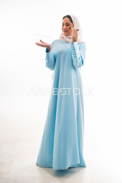 الأنوثة والاهتمام بالجمال والبشرة والمظهر الخارجي, مفهوم الدعاية والإعلان، خبيرة لمستحضرات التجميل النسائية، بورتريه لإمرأة عربية خليجية عمانية ترتدي العباءة والحجاب تقوم بوضع أحمر الخدود، التسويق لمنتجات الميك اب، خلفية بيضاء