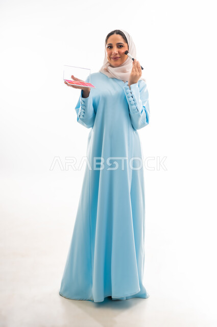 الأنوثة والاهتمام بالجمال والبشرة والمظهر الخارجي, مفهوم الدعاية والإعلان، خبيرة لمستحضرات التجميل النسائية، بورتريه لإمرأة عربية خليجية عمانية ترتدي العباءة والحجاب تقوم بوضع أحمر الخدود، التسويق لمنتجات الميك اب، خلفية بيضاء
