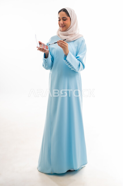 مفهوم الدعاية والتسويق، الأنوثة والاهتمام بالجمال والبشرة والمظهر الخارجي, خبيرة لمستحضرات التجميل النسائية، بورتريه مقرب لإمرأة عربية خليجية عمانية محجبة ترتدي عباءة زرقاء تقوم بوضع أحد مساحيق المكياج، التسويق لمنتجات الميك اب، خلفية بيضاء