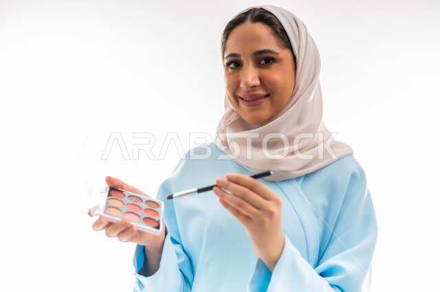 Advertising and marketing concept, femininity and interest in beauty, skin and appearance, female cosmetics expert, close-up portrait of a veiled Gulf Arab Omani woman wearing a blue abaya applying makeup, marketing for makeup products, white background