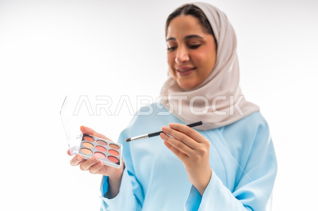 مفهوم الدعاية والتسويق، الأنوثة والاهتمام بالجمال والبشرة والمظهر الخارجي, خبيرة لمستحضرات التجميل النسائية، بورتريه مقرب لإمرأة عربية خليجية عمانية محجبة ترتدي عباءة زرقاء تقوم بوضع أحد مساحيق المكياج، التسويق لمنتجات الميك اب، خلفية بيضاء