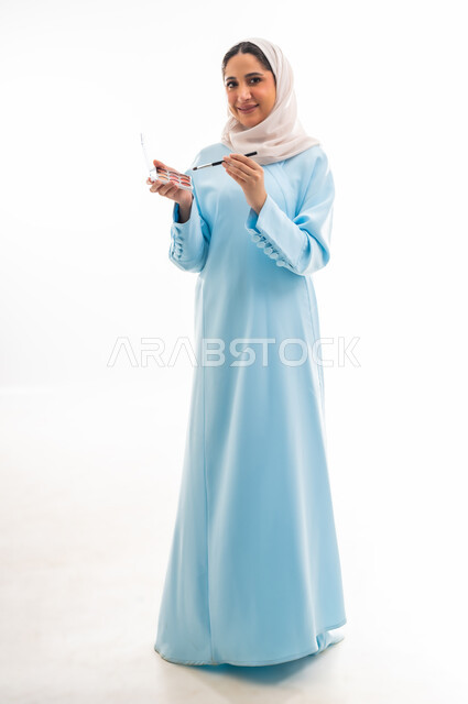 مفهوم الدعاية والتسويق، الأنوثة والاهتمام بالجمال والبشرة والمظهر الخارجي, خبيرة لمستحضرات التجميل النسائية، بورتريه مقرب لإمرأة عربية خليجية عمانية محجبة ترتدي عباءة زرقاء تقوم بوضع أحد مساحيق المكياج، التسويق لمنتجات الميك اب، خلفية بيضاء