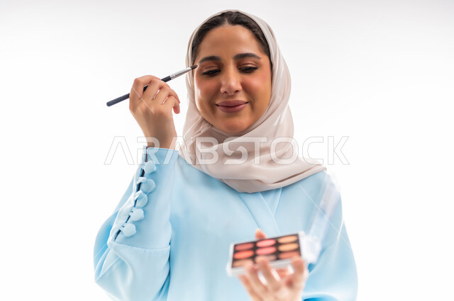 مفهوم الدعاية والتسويق، الأنوثة والاهتمام بالجمال والبشرة والمظهر الخارجي, خبيرة لمستحضرات التجميل النسائية، بورتريه مقرب لإمرأة عربية خليجية عمانية محجبة ترتدي عباءة زرقاء تقوم بوضع أحد مساحيق المكياج، التسويق لمنتجات الميك اب، خلفية بيضاء