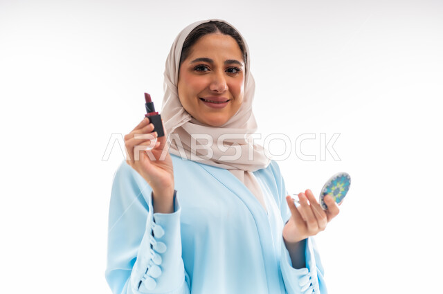 التسويق لمنتجات الميك اب، مفهوم الأنوثة والاهتمام بالجمال والبشرة والمظهر الخارجي, خبيرة لمستحضرات التجميل النسائية، بورتريه لإمرأة عربية خليجية عمانية ترتدي العباءة والحجاب  تحمل مرآة مدورة وقلم أحمر شفاه، خلفية بيضاء