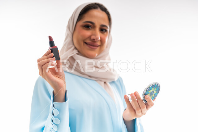 التسويق لمنتجات الميك اب، مفهوم الأنوثة والاهتمام بالجمال والبشرة والمظهر الخارجي, خبيرة لمستحضرات التجميل النسائية، بورتريه لإمرأة عربية خليجية عمانية ترتدي العباءة والحجاب  تحمل مرآة مدورة وقلم أحمر شفاه، خلفية بيضاء