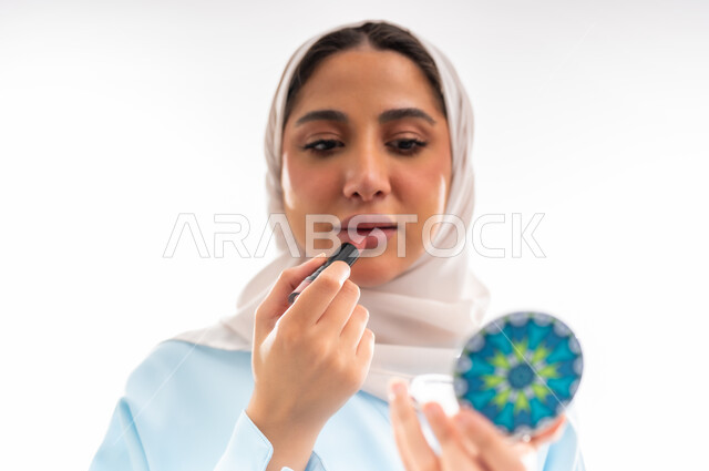 التسويق لمنتجات الميك اب، مفهوم الأنوثة والاهتمام بالجمال والبشرة والمظهر الخارجي, خبيرة  لمستحضرات التجميل النسائية، بورتريه لإمرأة عربية خليجية عمانية ترتدي العباءة والحجاب تقوم بوضع أحمر الشفاه، خلفية بيضاء