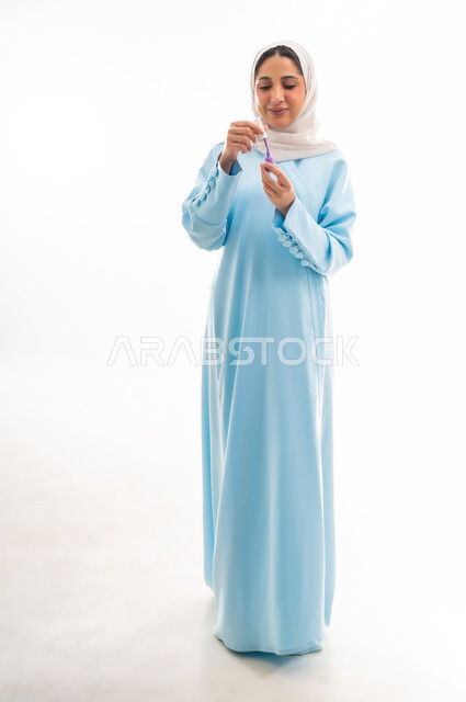 The concept of femininity and interest in beauty, skin and appearance, a female cosmetics expert, a close-up portrait of an Arab Gulf Omani woman wearing an abaya and hijab applying purple nail polish, marketing makeup products, white background