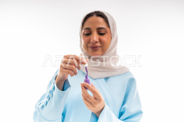 مفهوم الأنوثة والاهتمام بالجمال والبشرة والمظهر الخارجي, خبيرة لمستحضرات التجميل النسائية، بورتريه مقرب لإمرأة عربية خليجية عمانية ترتدي العباءة والحجاب تقوم بوضع طلاء الاظافر باللون البنفسجي، التسويق لمنتجات الميك اب، خلفية بيضاء