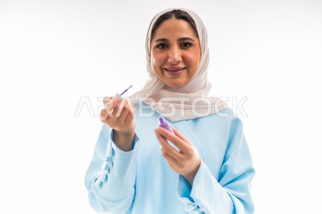مفهوم الأنوثة والاهتمام بالجمال والبشرة والمظهر الخارجي, خبيرة لمستحضرات التجميل النسائية، بورتريه مقرب لإمرأة عربية خليجية عمانية ترتدي العباءة والحجاب تقوم بوضع طلاء الاظافر باللون البنفسجي، التسويق لمنتجات الميك اب، خلفية بيضاء