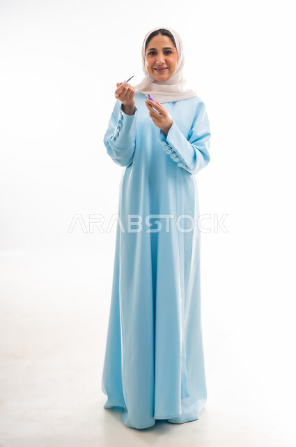The concept of femininity and interest in beauty, skin and appearance, a female cosmetics expert, a close-up portrait of an Arab Gulf Omani woman wearing an abaya and hijab applying purple nail polish, marketing makeup products, white background
