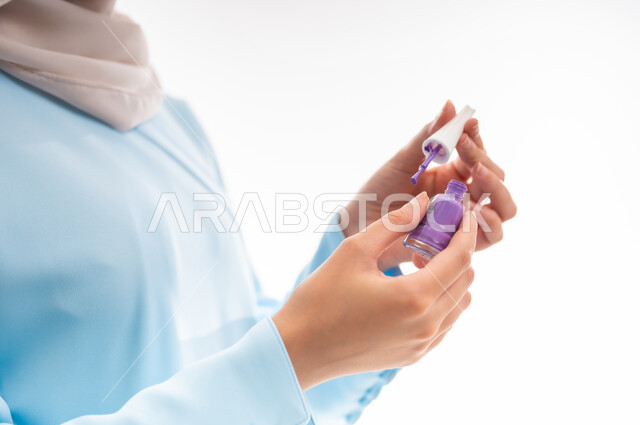 The concept of femininity and interest in beauty, skin and appearance, a female cosmetics expert, a close-up portrait of an Arab Gulf Omani woman wearing an abaya and hijab applying purple nail polish, marketing makeup products, white background
