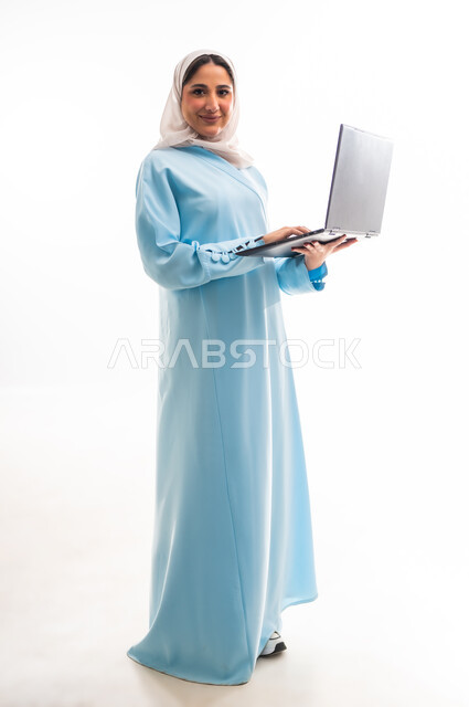 إجراء مكالمات فيديو عبر الانترنت، إنجاز مهام العمل عن بعد، مفهوم التقنيات والتكنولوجيا الحديثة المتطورة، بورتريه لامرأة عربية خليجية عمانية مبتسمة محجبة ترتدي عباءة زرقاء تقوم باستخدام جهاز الحاسوب المحمول، الجسم بالطول الكامل، خلفية بيضاء
