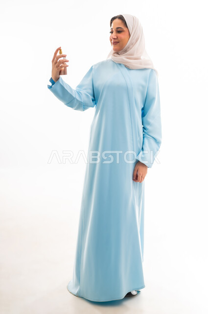 Marketing of national perfume products in Oman, using luxury perfumes, the concept of elegance and interest in one's appearance, using natural aromatic oils, portrait of a veiled Gulf Arab Omani woman wearing a blue abaya holding a bottle in her hand and spraying perfume, white background