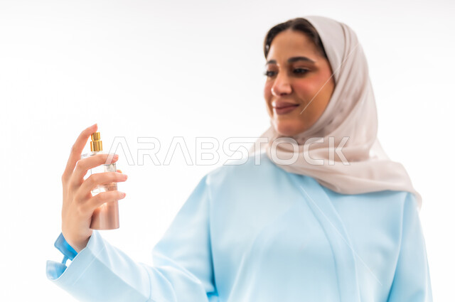 التسويق لمنتجات الروائح الوطنية في عمان، التطيب بالعطور الفاخرة، مفهوم الأناقة والاهتمام بالمظهر الخارجي، استخدام الزيوت العطرية الطبيعية، بورتريه لسيدة عربية خليجية عمانية محجبة ترتدي عباءة زرقاء تمسك بيدها زجاجة وتقوم برش العطر, خلفية بيضاء