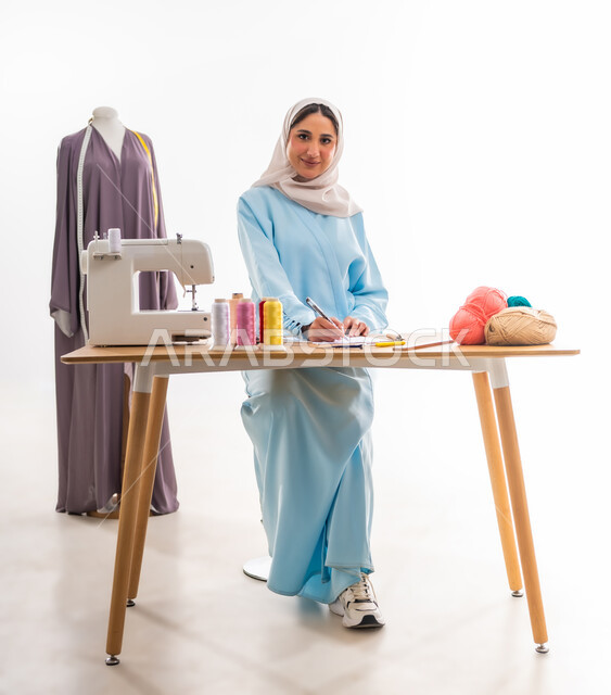 اختراع وابتكار تصاميم جديدة وعصرية، أهمية المرأة في مجال الخياطة، بورتريه لمصممة خليجية عمانية ترتدي عباءة زرقاء تقوم برسم تصميمات حديثة ومبتكرة، ورشة عمل تصميم ازياء، مهن وأعمال فنية نسائية، فنون ومهارات تفصيل الملابس النسائية، مفهوم عالم الأزياء والموضة
