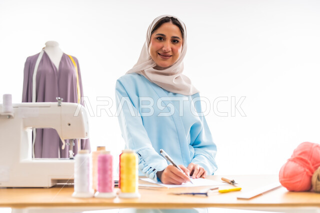 Inventing and creating new and modern designs, the importance of women in the field of sewing, a portrait of a Gulf Omani designer wearing a blue abaya drawing modern and innovative designs, a fashion design workshop, women’s professions and artistic works, arts and skills of tailoring women’s clothing, the concept of the world of fashion and style