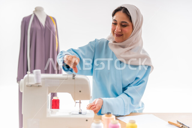 فنون ومهارات تفصيل الملابس النسائية، اختراع وابتكار تصاميم جديدة وعصرية، بورتريه لمصممة عربية خليجية عمانية ترتدي عباءة زرقاء تقوم بالعمل على آلة الحياكة، أهمية المرأة في مجال الخياطة، ورشة عمل تصميم ازياء، مهن وأعمال فنية نسائية، مفهوم عالم الأزياء والموضة