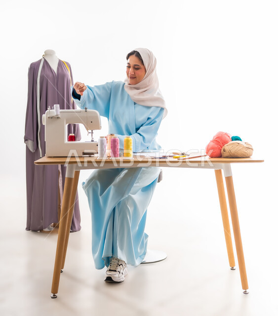 Arts and skills of tailoring women's clothing, invention and innovation of new and modern designs, portrait of an Arab Gulf Omani designer wearing a blue abaya working on a sewing machine, importance of women in the field of sewing, fashion design workshop, women's professions and artistic works, concept of the world of fashion and style
