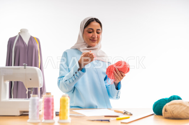 أهمية المرأة في مجال الخياطة، اختراع وابتكار موديلات جديدة وعصرية، بورتريه لامرأة عربية خليجية عمانية ترتدي عباءة زرقاء تحمل بيدها بكرة من خيوط الصوف، ورشة عمل تصميم ازياء، مهن وأعمال فنية نسائية، فنون ومهارات تفصيل الملابس النسائية، مفهوم عالم الأزياء والموضة