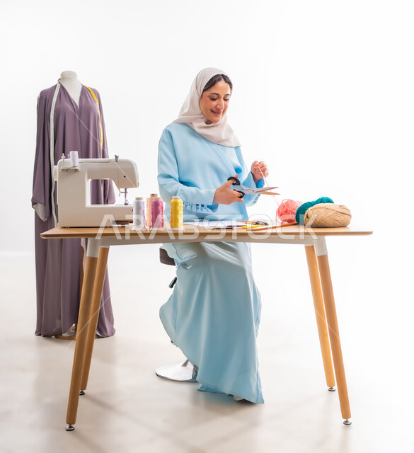 The concept of the world of fashion and style, invention and innovation of new and modern designs, portrait of an Arab Gulf Omani designer wearing a blue abaya cutting a woolen blanket, the importance of women in the field of sewing, fashion design workshop, women's professions and artistic works, arts and skills of tailoring women's clothing
