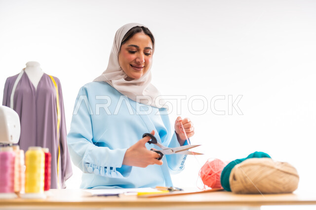 مفهوم عالم الأزياء والموضة، اختراع وابتكار تصاميم جديدة وعصرية، بورتريه لمصممة عربية خليجية عمانية ترتدي عباءة زرقاء تقوم بقص هيط من الصوف، أهمية المرأة في مجال الخياطة، ورشة عمل تصميم ازياء، مهن وأعمال فنية نسائية، فنون ومهارات تفصيل الملابس النسائية