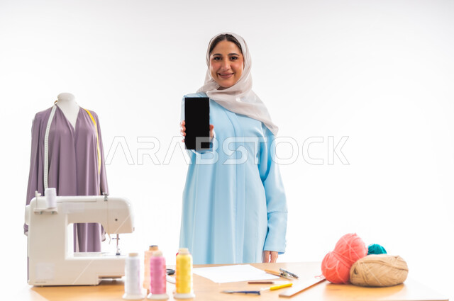 Arts and skills of tailoring women's clothing, invention and innovation of new and modern models, portrait of a smiling veiled Omani designer displaying a blank black phone screen, the importance of women in the field of sewing, fashion design workshop, women's professions and artistic works, the concept of the world of fashion and style