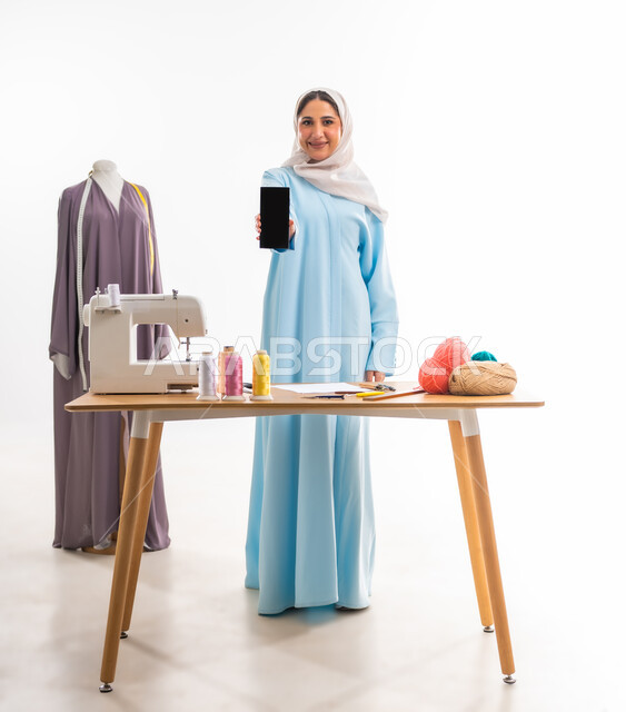 فنون ومهارات تفصيل الملابس النسائية، اختراع وابتكار موديلات جديدة وعصرية، بورتريه لمصممة عمانية محجبة مبتسمة تقوم بعرض شاشة هاتف سوداء فارغة أهمية المرأة في مجال الخياطة، ورشة عمل تصميم ازياء، مهن وأعمال فنية نسائية، مفهوم عالم الأزياء والموضة