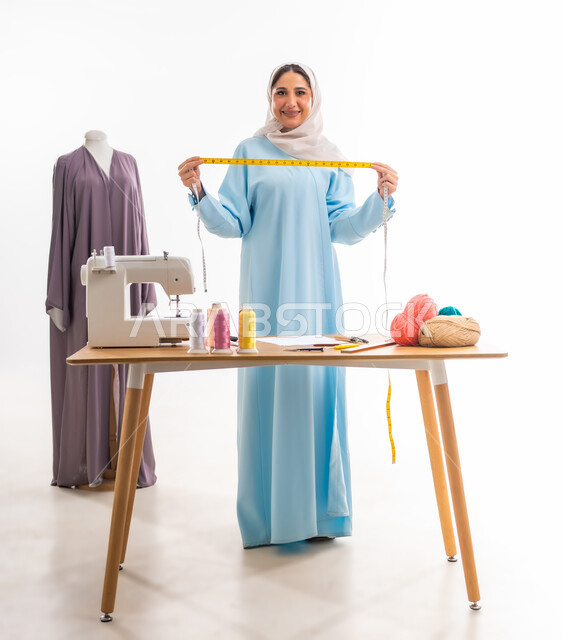 Women's professions and artworks, portrait of an Arab Gulf Omani designer holding a tape measure and looking at the camera with a smiling expression, invention and innovation of new and modern designs, the importance of women in the field of sewing, fashion design workshop, arts and skills of tailoring women's clothing, the concept of the world of fashion and style