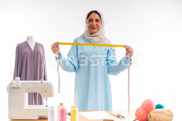 Women's professions and artworks, portrait of an Arab Gulf Omani designer holding a tape measure and looking at the camera with a smiling expression, invention and innovation of new and modern designs, the importance of women in the field of sewing, fashion design workshop, arts and skills of tailoring women's clothing, the concept of the world of fashion and style