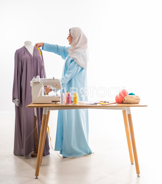 اختراع وابتكار تصاميم جديدة وعصرية، بورتريه لمرأة عربيه خليجيه عمانية ترتدي عباءة زرقاء تقوم بأخذ مقاسات ثوب،  أهمية المرأة في مجال الخياطة، ورشة عمل تصميم ازياء، مهن وأعمال فنية نسائية، فنون ومهارات تفصيل الملابس النسائية، مفهوم عالم الأزياء والموضة